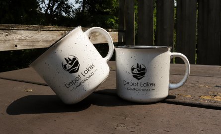 A photo of two white speckled mugs with the Quinte Conservation Depot Lakes Campground logo.